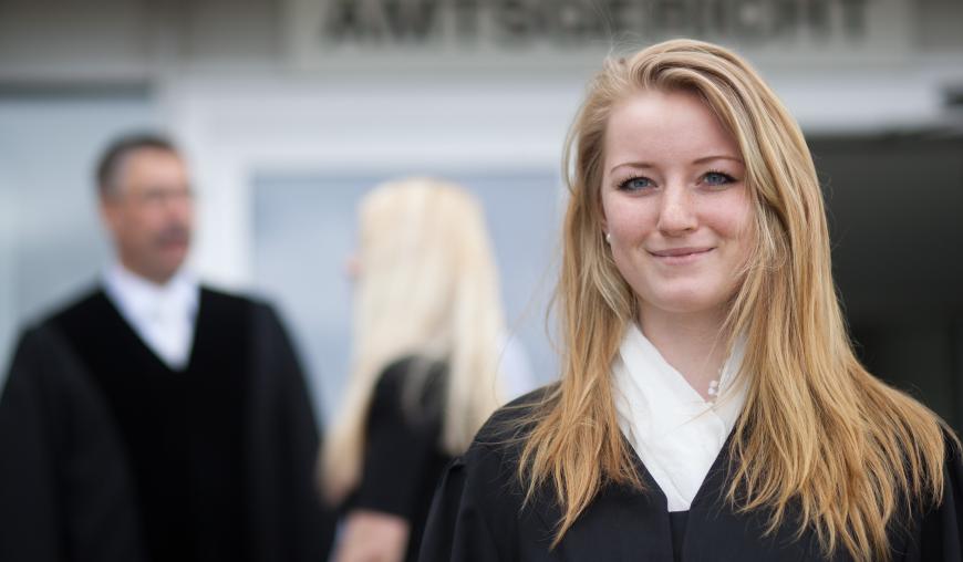 Im Vordergrund blickt eine Justizfachangestellte freundlich in die Kamera. Im Hintergrund unterhalten sich ein Staatsanwalt und eine Frau.