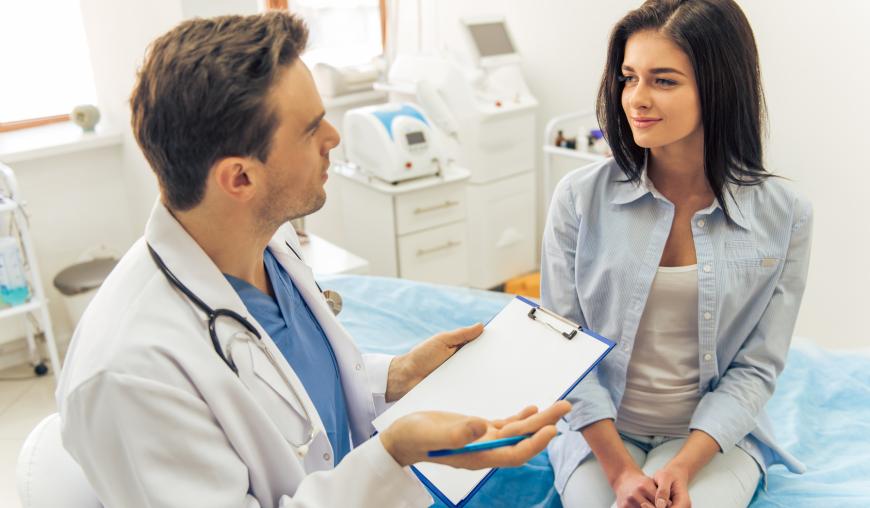 Ein Arzt spricht in seinem Behandlungszimmer zu einer Patientin, die auf einer Untersuchungsliege sitzt. In der Hand hält er ein Klemmbrett mit einen Anamnesebogen.