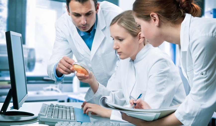 Eine Frau in weißem Kittel sitzt an einem Arbeitsplatz, vor einem Computer. In der Hand hält sie eine Petrischale, die ihr von einem Mann in weißem Kittel gereicht wurde. Eine weitere Frau in weißem Kittel steht daneben und macht sich Notizen.