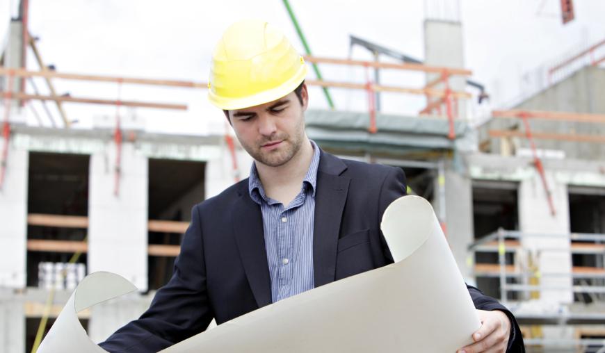Ein Mann mit gelbem Schutzhelm steht vor einer Rohbaustelle. In der Hand hält er eine Bauzeichnung.