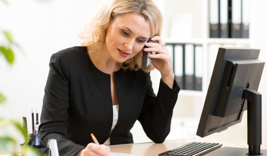 Eine Frau sitzt in einem Büro vor ihrem Computer. Sie telefoniert konzentriert und macht sich Notizen.