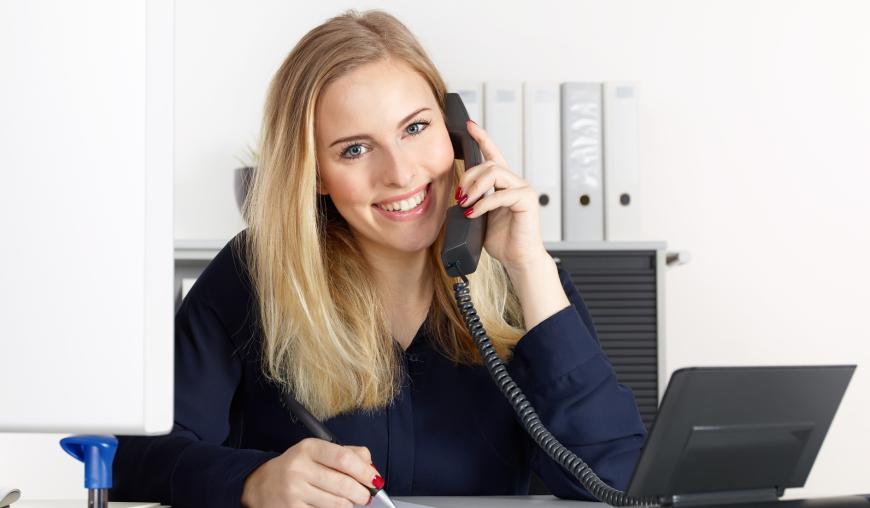 Eine Frau sitzt an einem Schreibtisch, vor einem Computer, telefoniert und macht sich nebenbei Notizen. Sie lächelt freundlich in die Kamera.