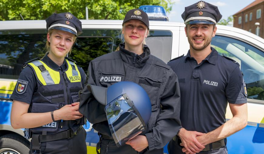 Drei Polizeifachkräfte – zwei weiblich, einer männlich – stehen in unterschiedlichen Uniformen und mit unterschiedlicher Ausstattung vor einem Polizeiwagen.