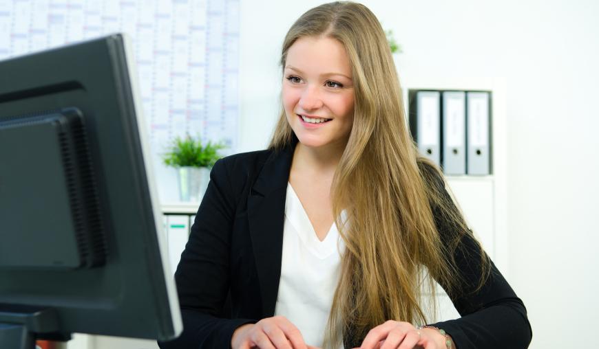 Eine Frau sitzt freundlich lächelnd am Schreibtisch, vor einem Computer. Sie tippt auf der Tastatur.