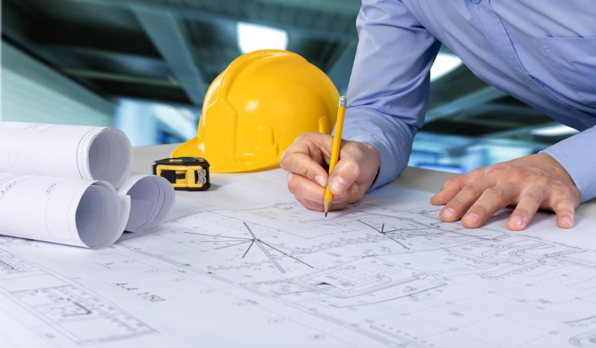 Ein Mann ist über einen Bauplan gebeugt und zeichnet etwas mit einem Bleistift ein. Neben dem Bauplan liegen weitere zusammengerollte Baupläne, ein Maßband und ein gelber Schutzhelm.