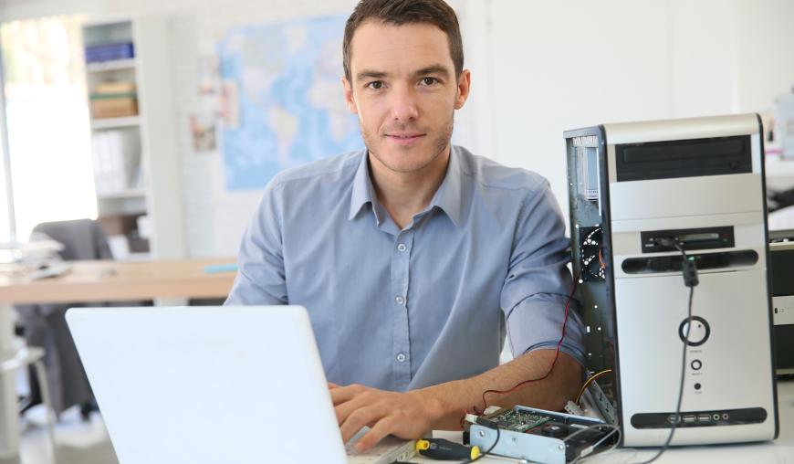 Ein IT-Fachmann sitzt mit einem Laptop in einem Büro. Das Laptop ist über ein Kabel mit einem PC verbunden, um diesen zu prüfen.