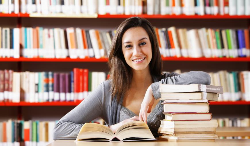 Eine Frau sitzt an einem Tisch, vor ihr ist ein Buch geöffnet. Mit einem Arm lehnt sie auf einem Stapel weiterer Bücher. Im Hintergrund steht ein volles Bücherregal.