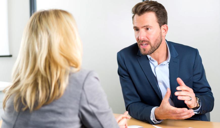 Eine Frau und ein Mann sitzen sich an einem Tisch gegenüber und diskutieren konzentriert. Zwischen ihnen liegen Unterlagen.