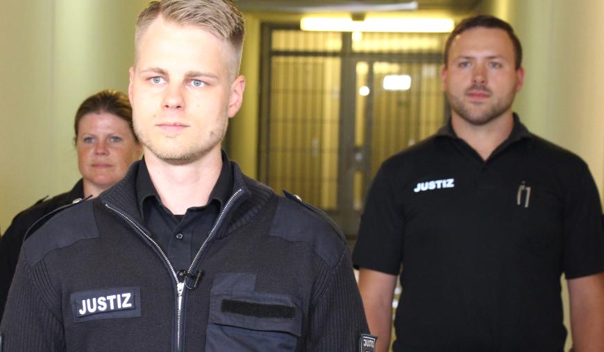 Zwei Justizvollzugsbeamte und eine Justizvollzugsbeamtin schreiten mit ernster Miene den Gefängnisgang entlang.