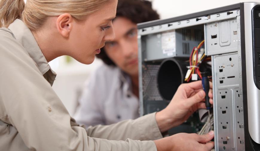 Eine Frau sitzt vor einem Computer mit geöffnetem Gehäuse und arbeitet an den Drähten des Geräts. Im Hintergrund sitzt ein Mann und schaut zu.