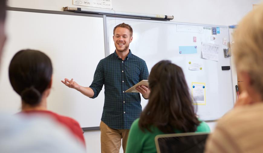 Ein Mann referiert mit einem freundlichen Lächeln vor interessierten Zuhörinnen und Zuhörern. Im Hintergrund hängt ein Whiteboard an der Wand, an dem Papiere befestigt sind.