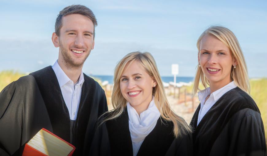 Zwei Frauen und ein Mann in Amtstracht und mit Gesetzestexten in den Händen blicken freundlich lächelnd in die Kamera.