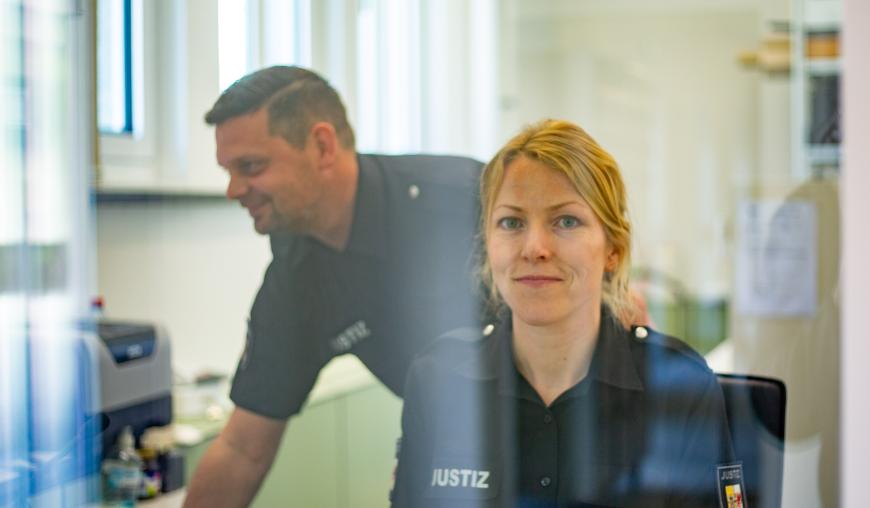 Eine Frau in Justiz-Uniform sitzt hinter einer Scheibe und blickt freundlich lächelnd in die Kamera. Im Hintergrund beugt sich ein Kollege Richtung Computer.