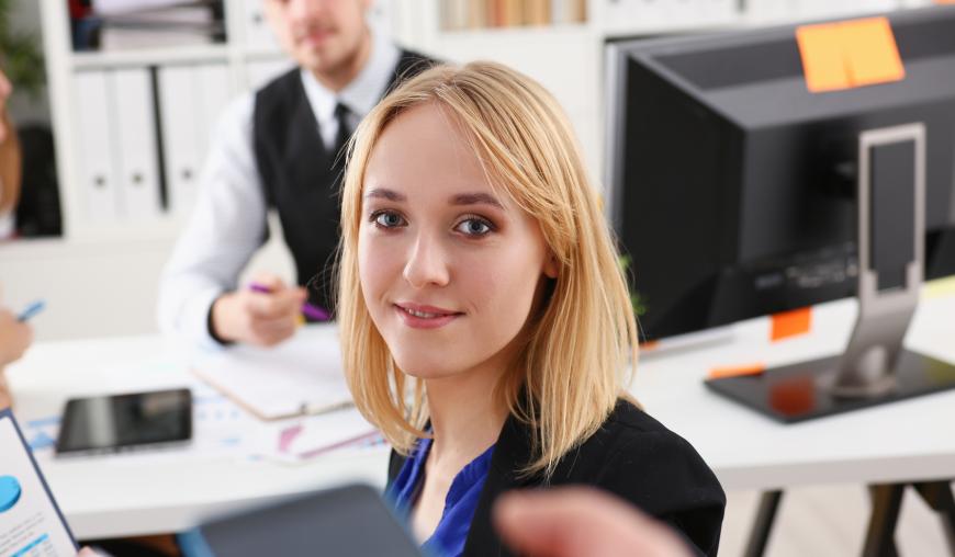 Eine Frau blickt freundlich in die Kamera, im Hintergrund sitzt ein Mann an einem Schreibtisch auf dem sich ein Computer-Bildschirm und Unterlagen befinden.