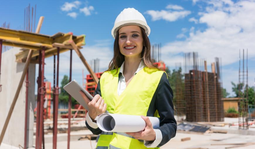 Eine Frau mit weißem Schutzhelm und gelber Warnschutzweste steht auf einer Baustelle. Im Hintergrund befinden sich unter anderem Stahlträger. In der Hand hält sie eine Bauzeichnung.