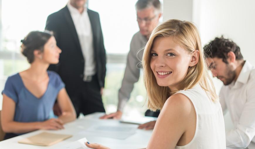 Drei Männer und zwei Frauen arbeiten konzentriert gemeinsam an einem Thema. Eine der Frauen blickt freundlich in die Kamera.