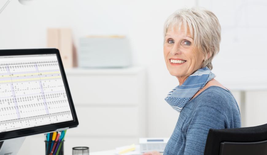 Eine freundlich lächelnde Frau in fortgeschrittenem Alter sitzt an einem Schreibtisch vor einem Computer und arbeitet an einer Excel-Tabelle.