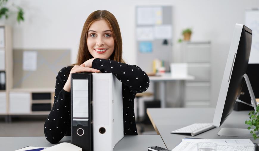 Eine freundlich lächelnde Frau sitzt in einem Büro und lehnt vor ihrem Computer auf einem weißen und einem schwarzen Aktenordner.