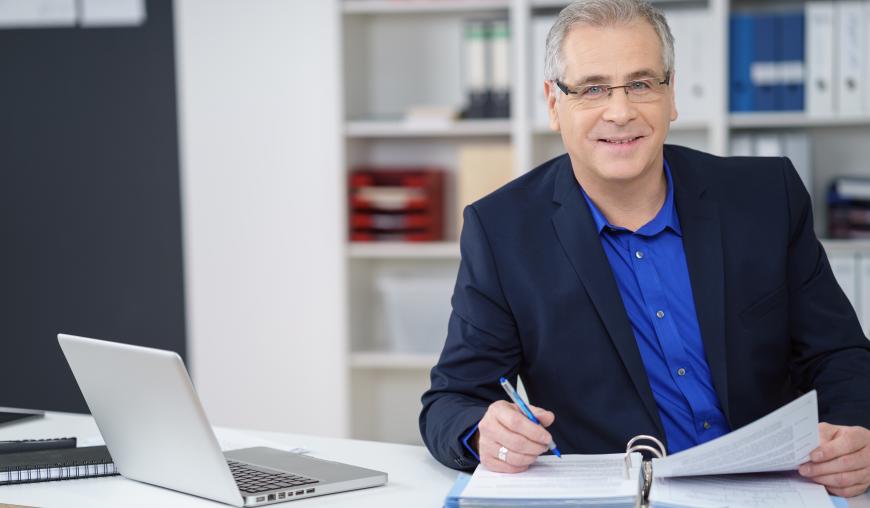 Ein Mann mittleren Alters sitzt an einem Schreibtisch und arbeitet Akten durch. Neben ihm steht ein Laptop.