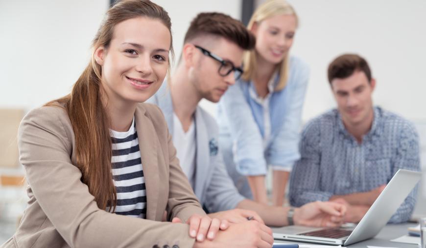 Zwei Frauen und zwei Männer sitzen und stehen gemeinsam um einen Laptop. Vor ihnen liegen Papiere. Eine Frau lächelt freundlich in die Kamera.