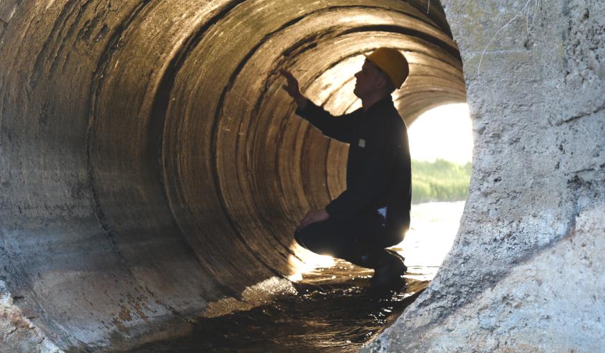 Ein Mann mit gelbem Schutzhelm und Arbeitsschutzkleidung hockt in einer Tunnelröhre und begutachtet die Beschaffenheit des massiven Rohres.