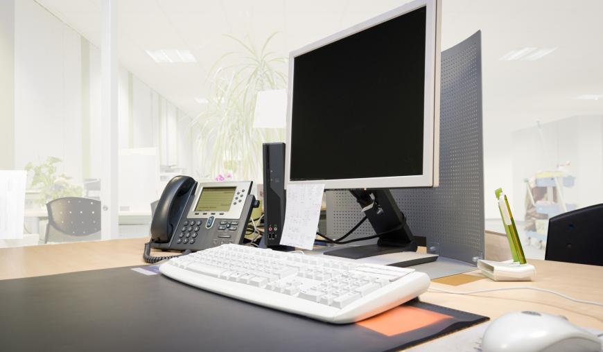 Ein Arbeitsplatz mit einem Computer, Bildschirm, einer Tastatur, einem Telefon und einer Maus. Im Hintergrund sind weitere Arbeitsplätze erkennbar.