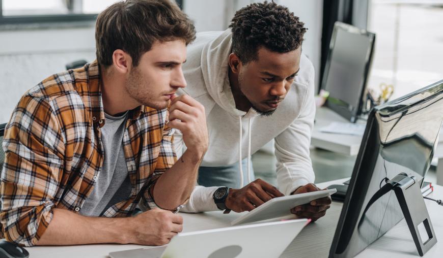 Zwei Männer mit unterschiedlicher Hautfarbe beugen sich gemeinsam über verschiedene IT-Technik und arbeiten konzentriert.