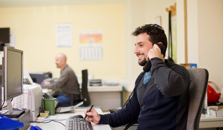 Ein Mann sitzt an einem Schreibtisch mit Computer in einem Großraumbüro und telefoniert.