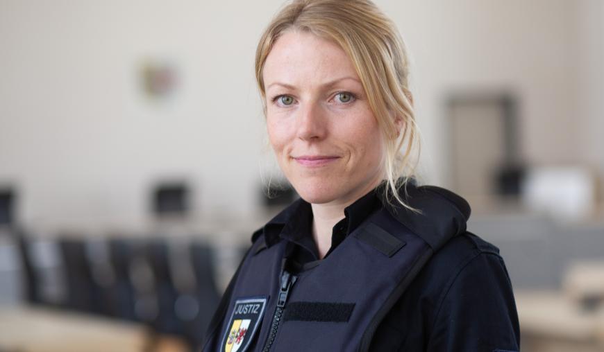 Eine Frau in Justiz-Uniform und mit einer Schutzweste blickt freundlich lächelnd in die Kamera. Im Hintergrund lässt sich ein Gerichtssaal erahnen.