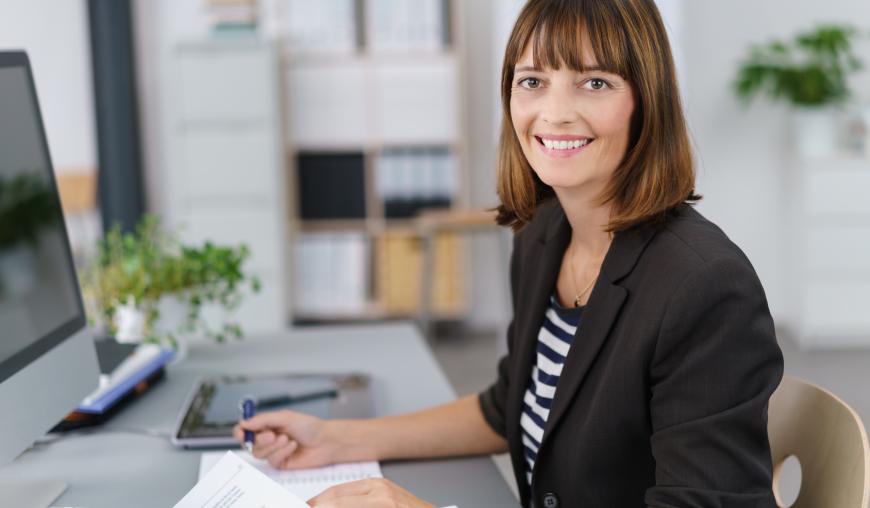 Eine Frau sitzt in einem Büro, an einem Schreibtisch, vor einem Computer und arbeitet an Unterlagen. Sie lächelt freundlich in die Kamera.