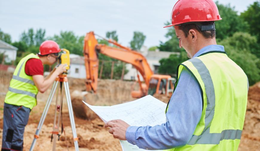 Zwei Männer mit roten Schutzhelmen und gelben Warnschutzwesten stehen auf einer Baustelle. Einer studiert eine Bauzeichnung, der andere schaut durch ein Messgerät. Im Hintergrund ist ein Bagger zugange.