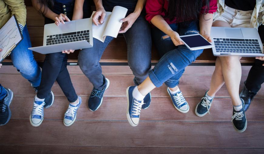 Eine Gruppe junger Menschen sitzt nebeneinander, auf ihrem Schoß halten sie Laptops, Tablets und Papierunterlagen. Sie tauschen sich aus.