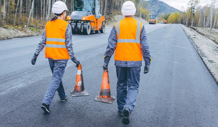 Eine Frau und ein Mann mit weißem Schutzhelm, orangefarbener Warnweste und Arbeitsschutzkleidung laufen auf einer frisch asphaltierten Straße. Sie tragen Sicherheitskegel. Im Hintergrund sind Baumaschinen.